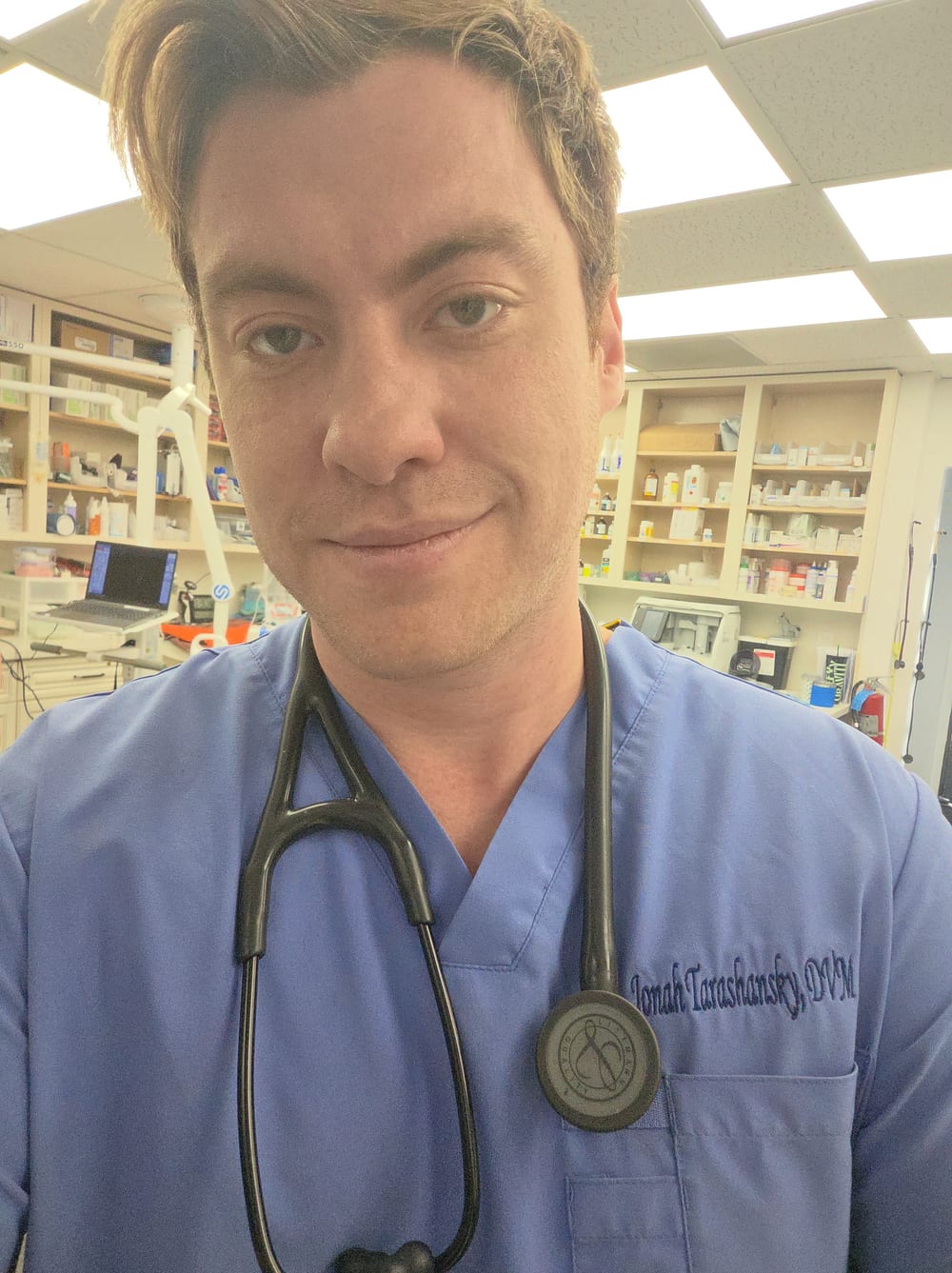 Jonah in blue scrubs embroidered “Jonah Tarashansky, DVM”, with a stethoscope around his neck, inside a veterinary clinic.